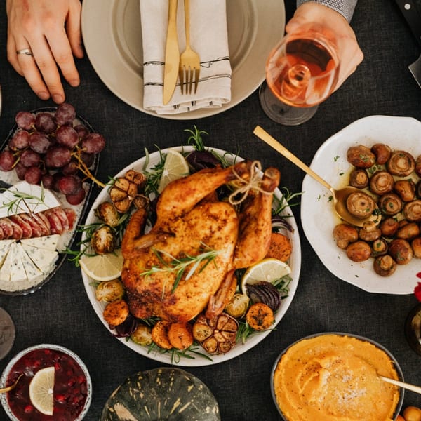 Overhead view of a roasted chicken dinner with sides, wine, and a person's hands.