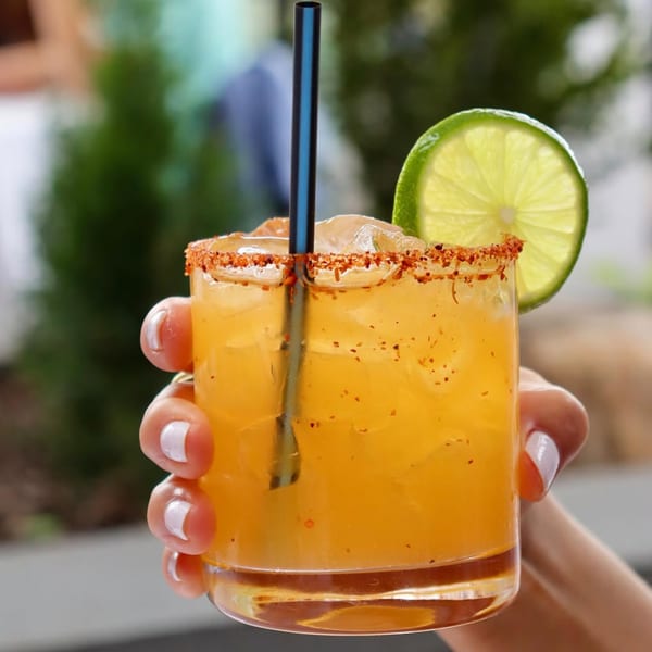 Hand holding an orange cocktail with a chili-lime rim, a black straw, and a lime slice.