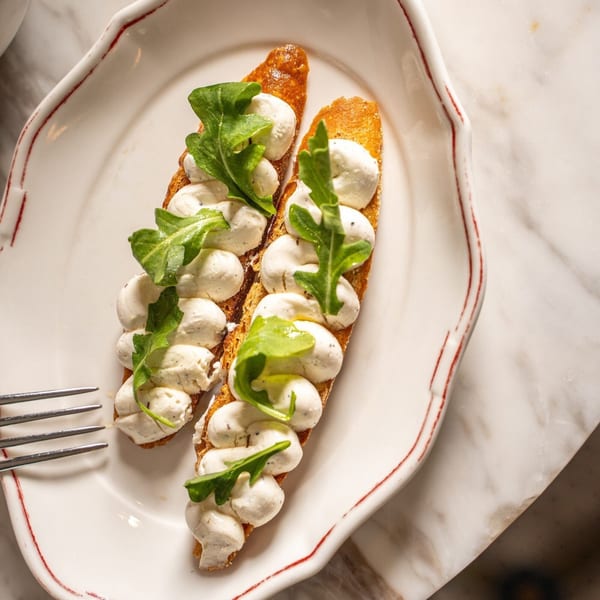 Two Italian crostini topped with creamy white cheese and fresh arugula on a white plate.