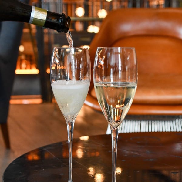 Sparkling wine being poured into a flute glass next to another full glass on a dark table.