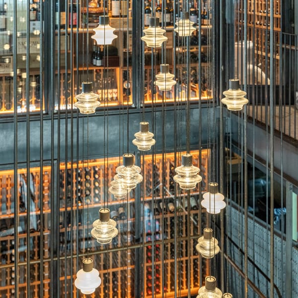 Hanging glass light fixtures illuminate a multi-story wine cellar with bottles visible on shelves.