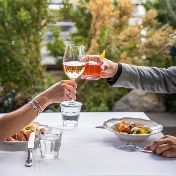 Two people clinking glasses of rosé and a cocktail at an outdoor table with Italian dishes.