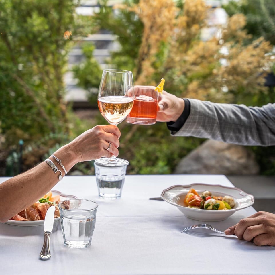 Two people clinking glasses of rosé and a cocktail at an outdoor table with Italian dishes.