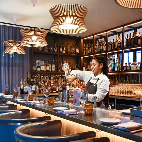 A smiling Officina bartender pours a drink at a well-stocked bar with stylish lighting and seating.