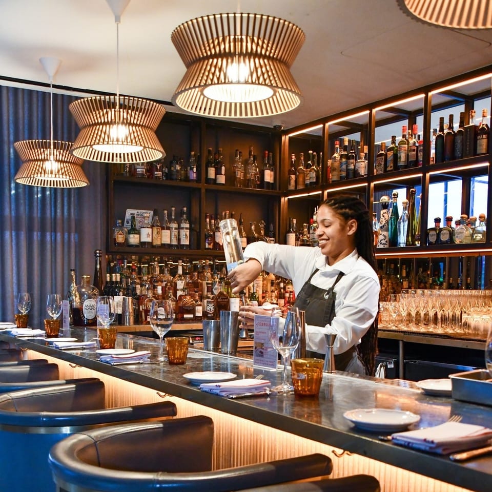 A smiling Officina bartender pours a drink at a well-stocked bar with stylish lighting and seating.