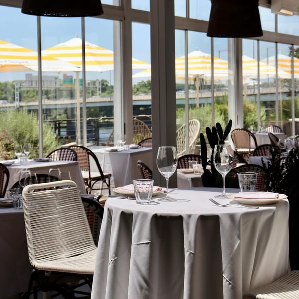 An Officina dining room with white-clothed tables, wicker chairs, and a view of outdoor umbrellas.