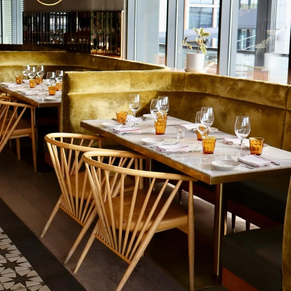 Officina restaurant interior with set tables, golden velvet banquettes, and wooden chairs by large windows.