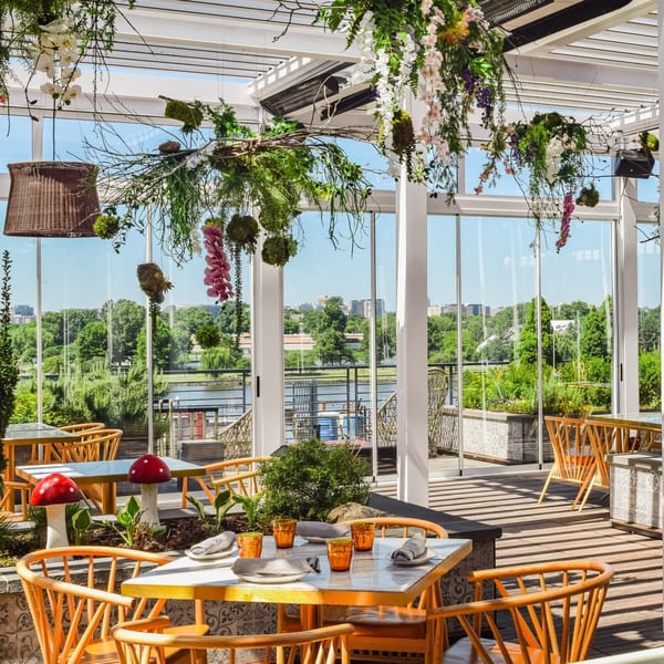 Officina's outdoor dining area with wooden chairs, set tables, and hanging plants overlooking a river.